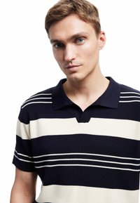 Polo w paski w odcieniach granatu i kremu, z teksturą prążkowaną, krótkimi rękawami oraz kołnierzykiem w kształcie V. Pionowe paski są wyraźnie widoczne.