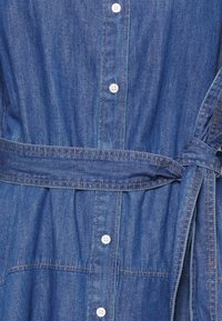 Blå denimklänning med krage, knapplist och bälte i midjan. Har fickor och synliga sömnadsdetaljer.