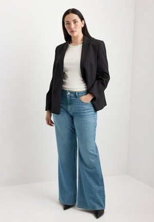 Žena stojí v čiernom blejzri, bielom svetri, modrých džínsach s širokými nohavicami a čiernych špicatých topánkach na jednoliatom bielom pozadí.