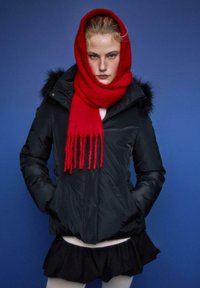 Črna podložena jakna z obrobo iz krzna, rdeč volnen šal, plastičast bel pulover in črna krila. Na modrem ozadju.