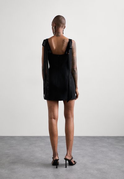 Vestido mini negro con un diseño ajustado, que muestra tirantes de hombro atados con cintas y una espalda baja. La modelo lleva sandalias de tacón alto negras.