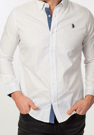 Homme portant une chemise blanche à manches longues avec un motif, patte de boutonnage intérieure bleue et pantalon noir, mains dans les poches.