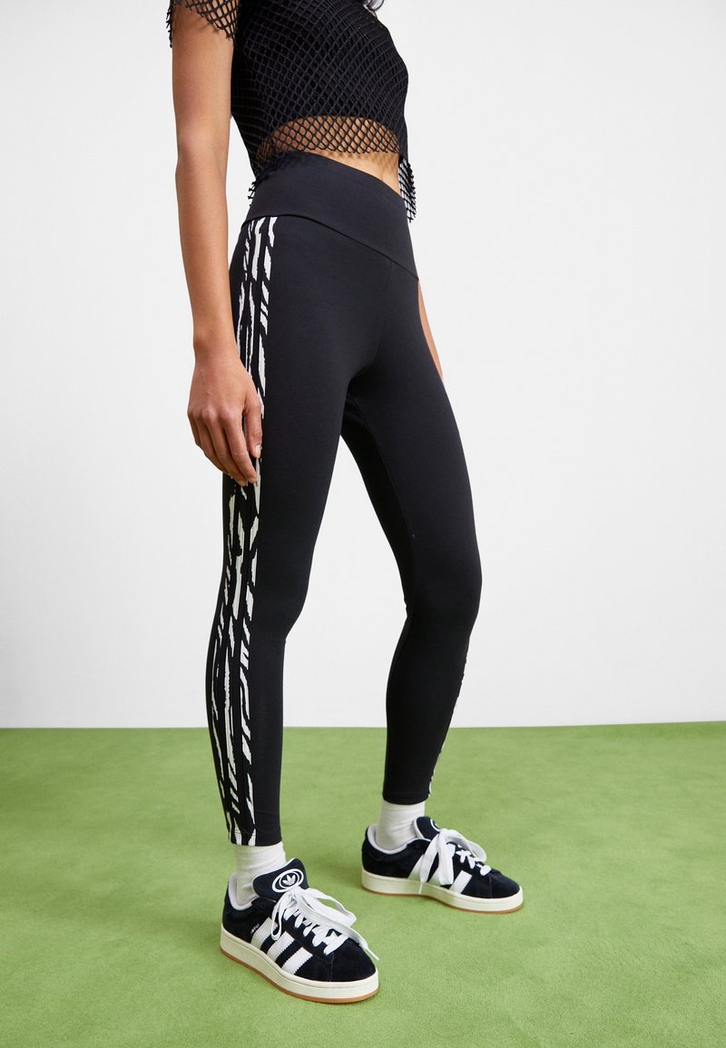 Leggings neri con vita alta, caratterizzati da un motivo a zigzag bianco lungo i lati, abbinati a scarpe da ginnastica nere e calze bianche.