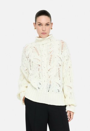 Donna con capelli scuri in un maglione a collo alto bianco con dettagli in perle e pantaloni neri, in piedi contro uno sfondo chiaro e neutro.