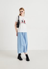 T-shirt bianca con un design a fumetti rosso e nero, abbinata a una lunga gonna maxi blu chiaro e stivali neri con lacci. Borsa nera, occhiali da sole.