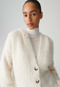 Weicher cremefarbener Cardigan mit tiefem V-Ausschnitt, strukturiertem Material, drei großen Knöpfen und weichen Kanten, getragen über einem leichten Rollkragenpullover.