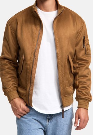 Homme portant une veste marron clair à fermeture éclair sur un t-shirt blanc et un jean bleu, debout avec une main dans la poche sur fond blanc.