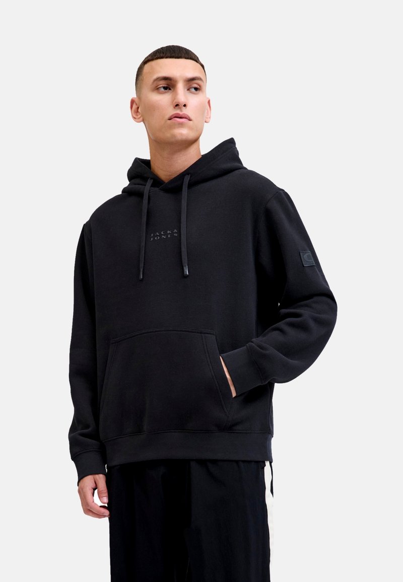 Schwarzer Kapuzenpullover mit Kängurutasche, strukturiertem Stoff, Logo auf der Brust, verstellbaren Kordeln und gerippten Bündchen. Modell trägt schwarze Hose.