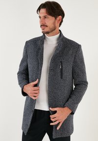 Cappotto strutturato blu scuro e bianco con motivi, colletto alto e tasca con zip. Maniche lunghe e taglio dritto, abbinato a un dolcevita chiaro.