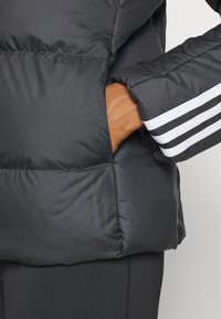 Giacca a vento nera con finitura opaca, dotata di tasca laterale e disegno a tre strisce bianche sulla manica. Texture liscia, materiale isolante.