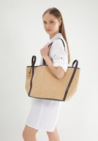 Stribet tote taske med mørkebrune læderdetaljer, rektangulær form, to håndtag og et rundt guld hardware logo fastgjort.