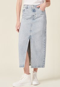 Gonna lunga in denim azzurro chiaro con spacco frontale, vita alta, cinque tasche e chiusura classica con bottone e cerniera. Indossata con sneaker bianche alte.