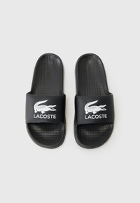 Black rubber slides with a wide band featuring a white crocodile logo and "LACOSTE" text. Textured footbed for grip and comfort.