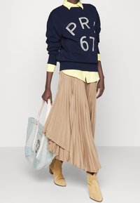 Navy sweatshirt with "PR" and "67" printed in white, layered over a light yellow shirt. Tan pleated skirt, beige ankle boots, and a light blue bag.