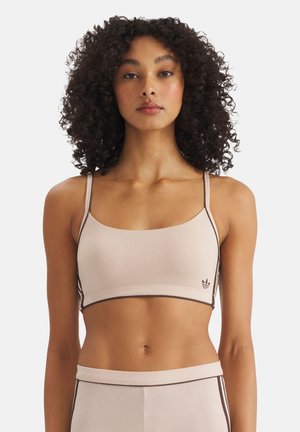 Žena s kudrnatými vlasy, oblékající béžový sportovní top a ladící legíny s tmavými lemy, stojící před jednobarevným bílým pozadím.