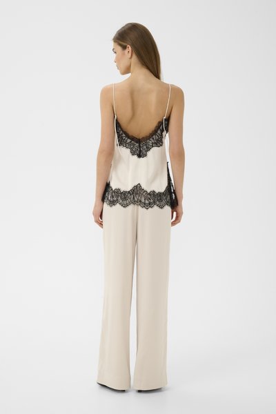 Femme debout de dos portant une camisole crème avec des détails en dentelle noire et un pantalon large crème assorti sur un fond uni.