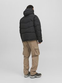 Giacca a vento nera con cappuccio, caratterizzata da un design trapuntato. Abbinata a pantaloni cargo beige e scarpe da ginnastica grigie, vista da dietro.