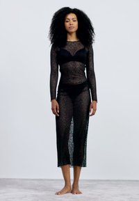 Conjunto em malha preta transparente com mangas longas e saia até ao meio da panturrilha. Padrão geométrico de diamante, combinado com um bralette e biquíni.