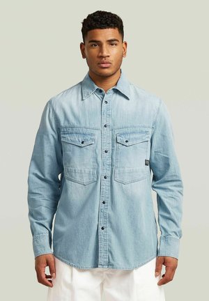 Junger Mann mit kurzen lockigen Haaren, der ein hellblaues Jeanshemd mit Knöpfen und weiße Hosen trägt, steht vor einem schlichten Hintergrund.