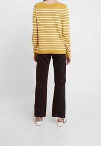 Maglione a righe gialle e bianche in maglia con scollatura ampia, abbinato a pantaloni a gamba larga marrone scuro. Sandali chiari completano l'outfit.