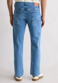 Osoba w niebieskich dżinsach o prostym kroju z tylnymi kieszeniami i białych sneakersach z zielonymi akcentami, stojąca przy gładkiej białej ścianie.