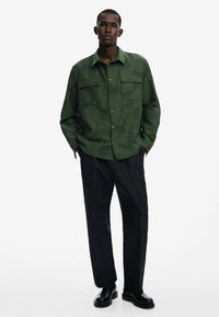 Green camo shirt with two chest pockets, button front, and long sleeves, paired with black trousers and smooth black shoes.