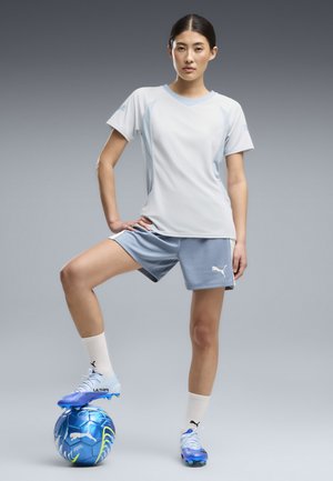 Una mujer luce una camiseta deportiva blanca, pantalones cortos azules y botas de fútbol azules, con un pie apoyado en un balón de fútbol azul sobre un fondo gris.