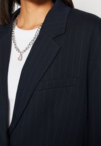 Persona che indossa un blazer blu a righe sopra una maglietta bianca a coste, con una collana d'argento a catena spessa che presenta piccoli ciondoli.