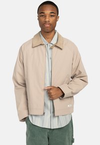 Chaqueta beige con cuello de ante, cierre de cremallera en la parte delantera y bolsillos laterales, llevada sobre una camisa a rayas azul y blanca. Textura suave y corte relajado.