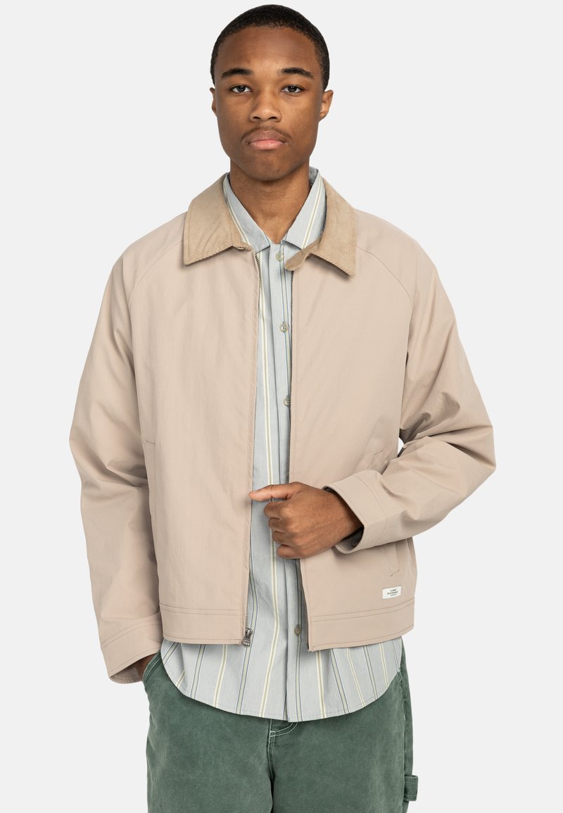 Chaqueta beige con cuello de ante, cierre de cremallera en la parte delantera y bolsillos laterales, llevada sobre una camisa a rayas azul y blanca. Textura suave y corte relajado.