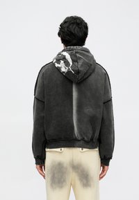 Sweatshirt cinzenta com capuz e desenho desbotado, apresentando um capuz grande e tecido texturizado. Combinada com calças de cor clara exibindo um padrão em gradiente.