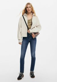 Skuren ljusbeige hoodie, olivgrön ribbad linne, mörkblå skinny jeans och svarta sneakers med grå snören; håller en mönstrad crossover-väska.