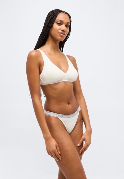 Γυναίκα με μακριά πλεξούδα που φοράει λευκό τριγωνικό σουτιέν και ασορτί εσώρουχο με γκρι ζώνη μέσης με λογότυπο Emporio Armani, στέκεται μπροστά σε λευκό φόντο.