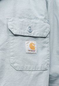 Τσέπη από ανοιχτό γκρι ύφασμα με καπάκι με κουμπί και ένα τετράγωνο έμβλημα λογότυπου Carhartt ραμμένο κάτω από το κέντρο του καπακιού.