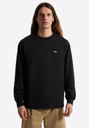 Junger Mann mit langen braunen Haaren trägt ein schwarzes Vans Langarmshirt und beige Hose, steht vor weißem Hintergrund.