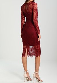 Burgundy lace dress with long sleeves and a knee-length hem, featuring a sheer back and intricate floral patterns. Paired with beige heels.