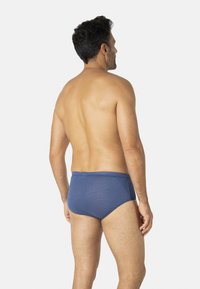 Slip de bain bleu texturé à taille basse, avec une ceinture lisse et un tissu à motif, porté par un mannequin masculin vu de dos.