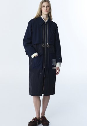 Kobieta w długim granatowym płaszczu z wieloma suwakami, przednimi kieszeniami i naszywkami, w połączeniu z brązowymi butami w stylu mokasynów z frędzlami.