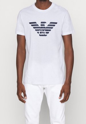 Mężczyzna w białym t-shircie z krótkim rękawem, z dużym czarnym logo orła GA na piersi oraz w białych spodniach, stoi na tle jednolitego tła.