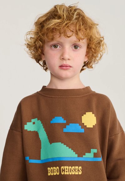 Enfant aux cheveux roux bouclés portant un sweat-shirt marron avec un dinosaure vert pixellisé, des nuages bleus, un soleil jaune et le texte "BOBO CHOSES".