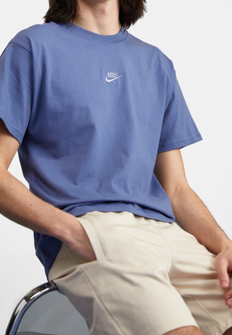 Camiseta de algodón azul con mangas cortas y cuello redondo, que presenta un pequeño logo blanco de Nike en el pecho. Lleva puesta un pantalón corto de color beige claro.