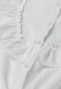 Tessuto bianco con un motivo a pois testurizzato. Presenta piccole balze lungo il colletto e bottoni trasparenti con accenti arancioni.