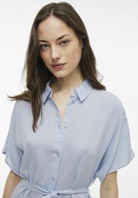 Camicia azzurro chiaro con colletto, maniche corte, apertura con bottoni e dettaglio in vita annodato. Tessuto liscio con una leggera lucentezza.