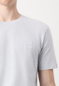 Svetlo siva bombažna majica s krojnim ovratnikom, kratkimi rokavi in majhnim, vtisnjenim kvadratnim logotipom na prsih. Mehka tekstura, preprost dizajn.