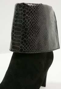 Botines negros con un puño de imitación de serpiente texturizado y brillante. El cuerpo está hecho de ante suave, con una forma elegante y en forma de cono y un tacón medio.