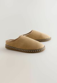 Tan slippers made of soft material with a smooth texture. They feature a braided accent along the sole and an open back design.
