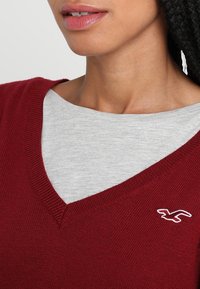 Burgunderfärgad stickad tröja med v-ringning, lagd över en ljusgrå t-shirt. Har en liten vit logotyp på vänster sida. Slät textur.