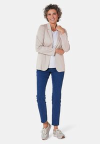 Lichtroze blazer over een witte shirt, gecombineerd met blauwe jeans en lichtgrijze sneakers. Kruidig haar, staande pose, ontspannen pasvorm.