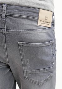 Jeans de mezclilla gris con un bolsillo trasero ligeramente desgastado, una etiqueta de marca de cuero beige y detalles de costura estándar.