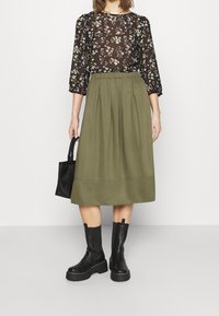 Svart blommig blus med 3/4-ärmar, olivgrön veckad midikjol, svarta platå ankelskors och en liten svart handväska.